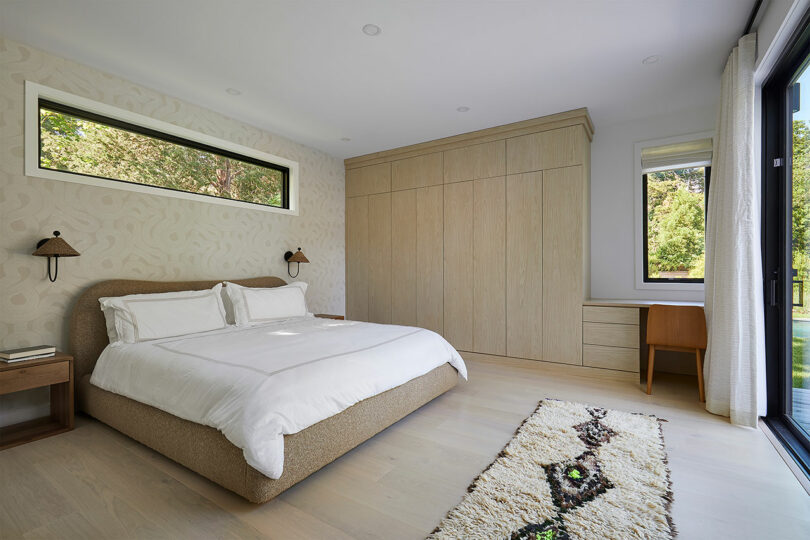 The Shift House-inspired bedroom features a modern aesthetic with a large bed, light wood flooring, and a built-in wardrobe. Natural light floods in through two windows, while a cozy rug is placed beside the bed for added comfort.