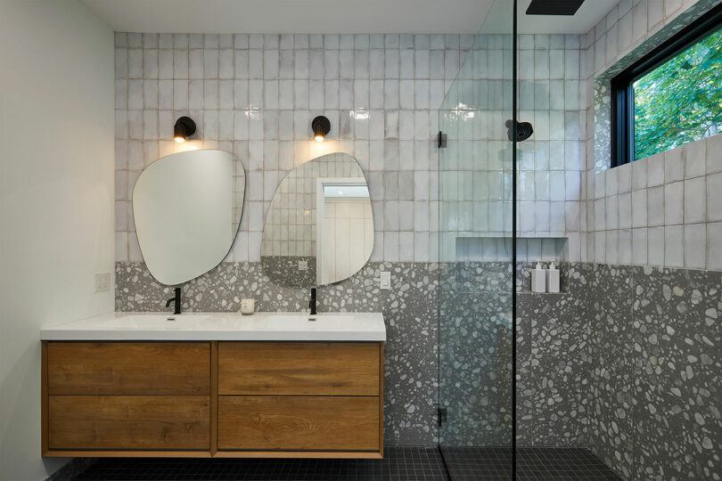The Shift House features a modern bathroom complete with a double sink vanity, elegant wood cabinets, and irregular mirrors. Tiled walls complement the glass-enclosed shower, while ample natural light pours through a rectangular window.