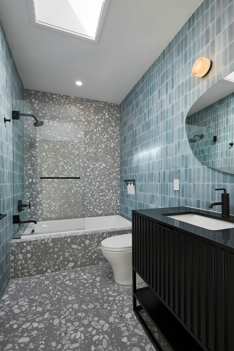 The modern bathroom at Shift House features blue tiled walls, a terrazzo floor, and a sleek bathtub. Black fixtures complement the elegant design, while a light above the mirror and a skylight on the ceiling enhance the airy atmosphere.