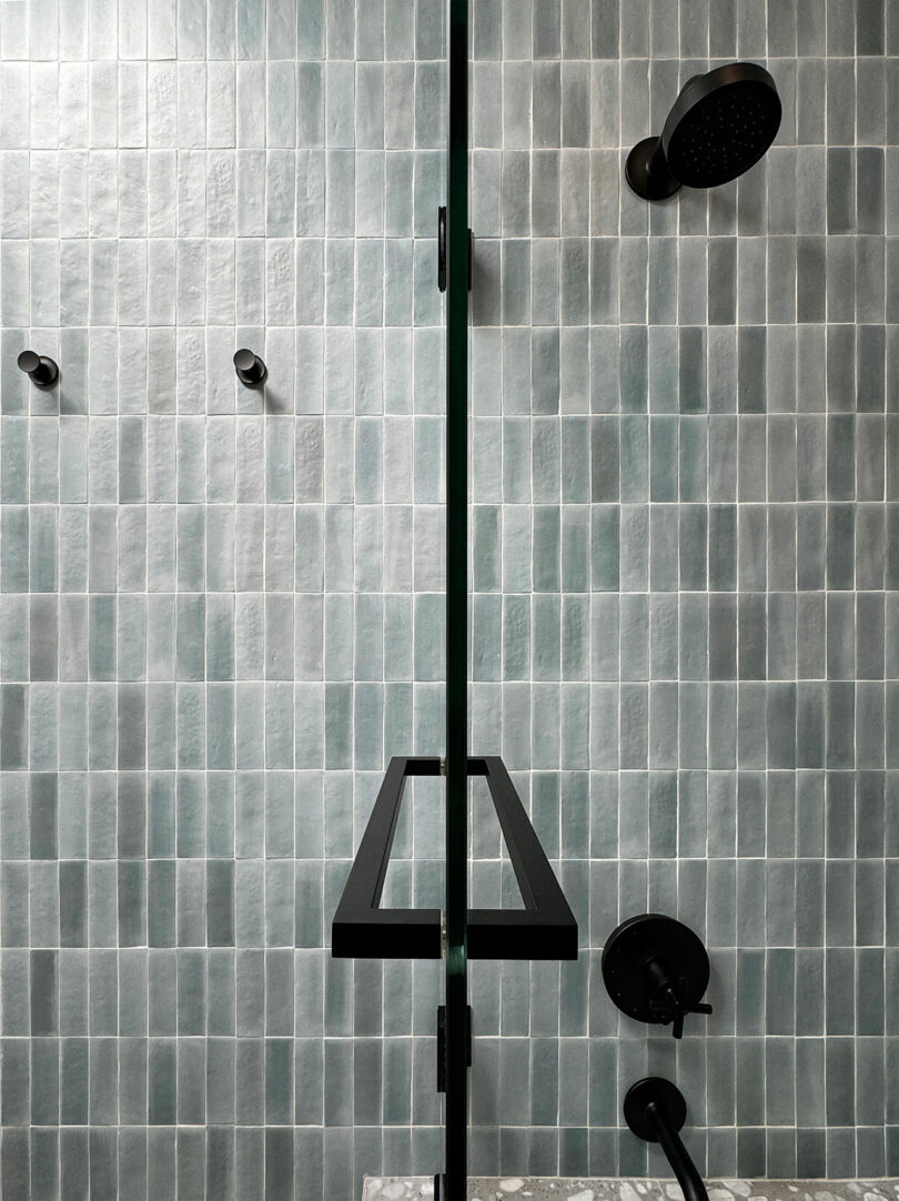A modern shower in Shift House features glass doors, a rainfall showerhead, and elegantly greenish-gray tiled walls.