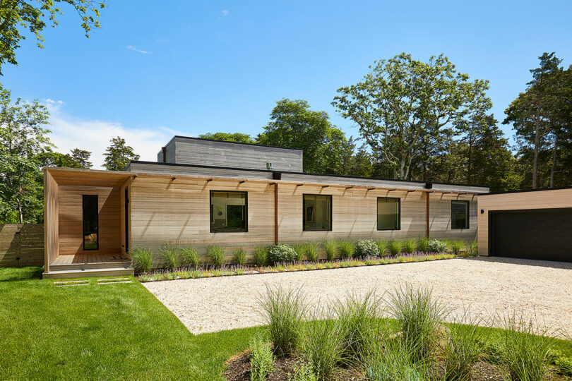 The Shift House is a modern single-story home featuring a sleek wooden exterior, flat roof, and expansive windows. A gravel driveway leads to the neatly landscaped garden with lush grass and shrubs, all nestled amid towering trees.