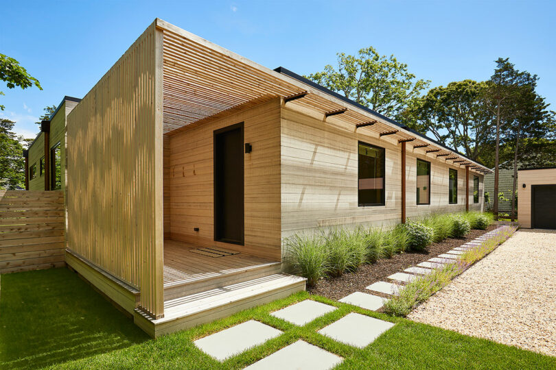 Shift House is a modern flat-roofed home featuring elegant wood paneling, a covered wooden porch, and a beautifully landscaped garden. A stone path meanders through the greenery, while the gravel driveway adds to its serene charm.