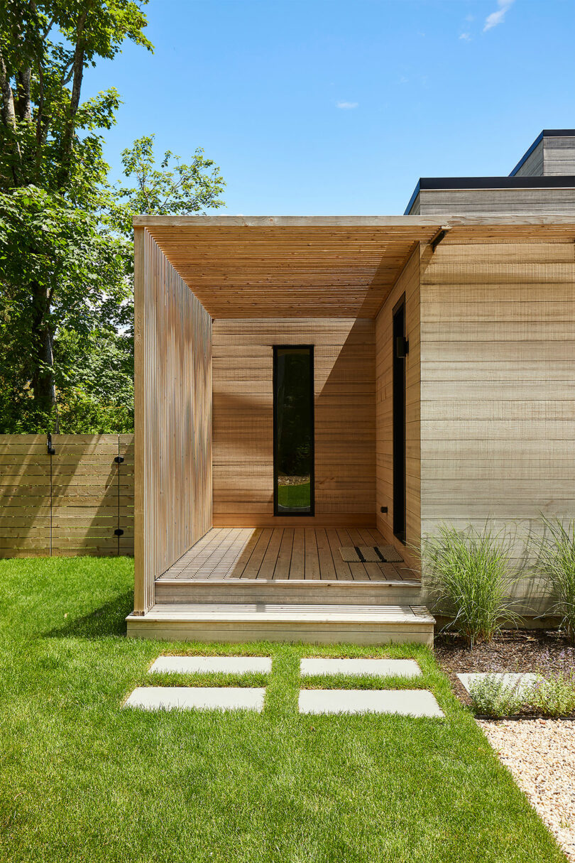 A modern wooden Shift House exterior with a small porch complements the yard's green grass and stepping stones leading to the entrance. Trees and a rustic wooden fence create a serene backdrop.