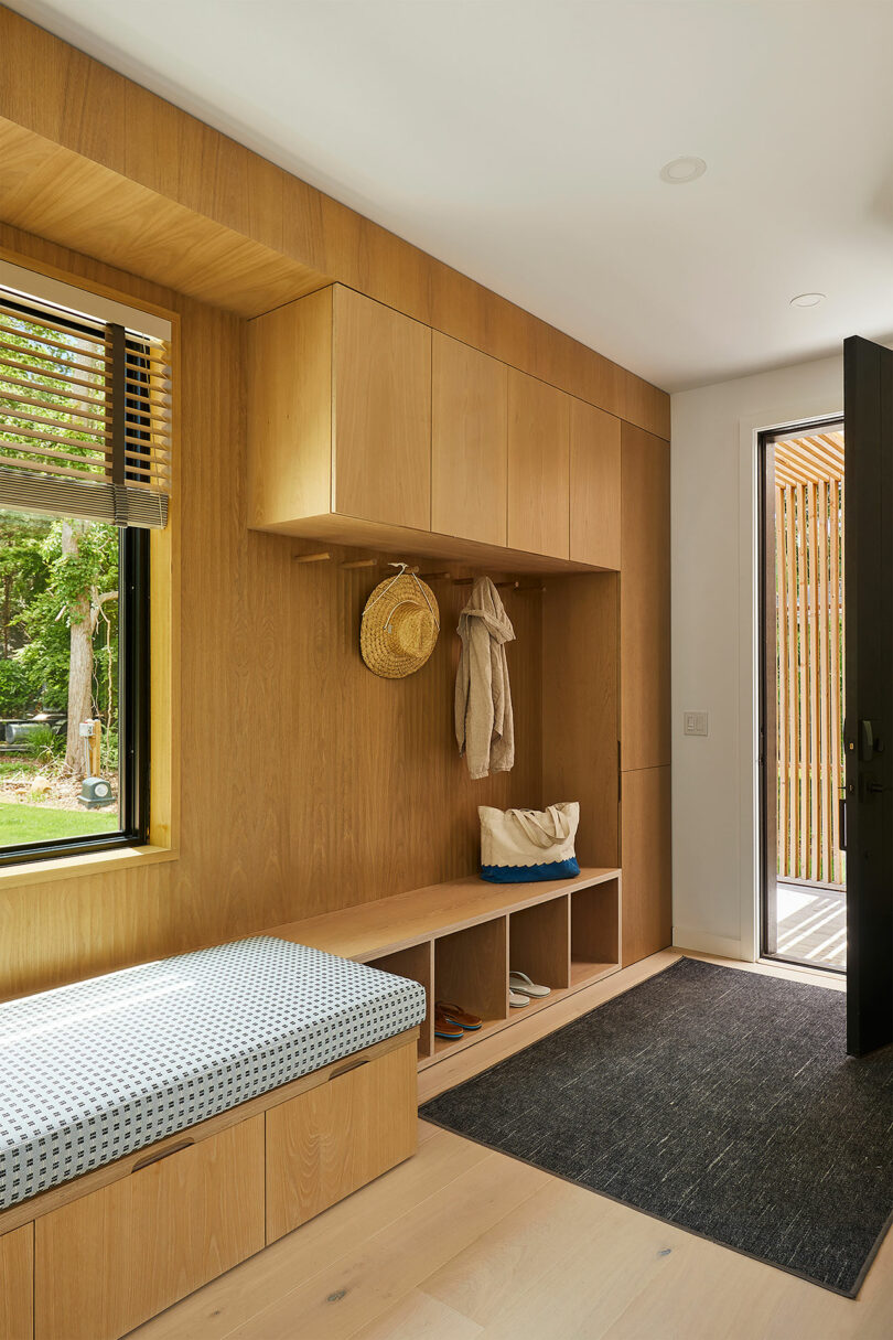 In this modern mudroom of the Shift House, wooden cabinetry complements a bench with a gray cushion. Wall hooks hold hats and bags above a stylish rug. A window with blinds allows soft light to filter in, while the door to the outside sits conveniently on the right.