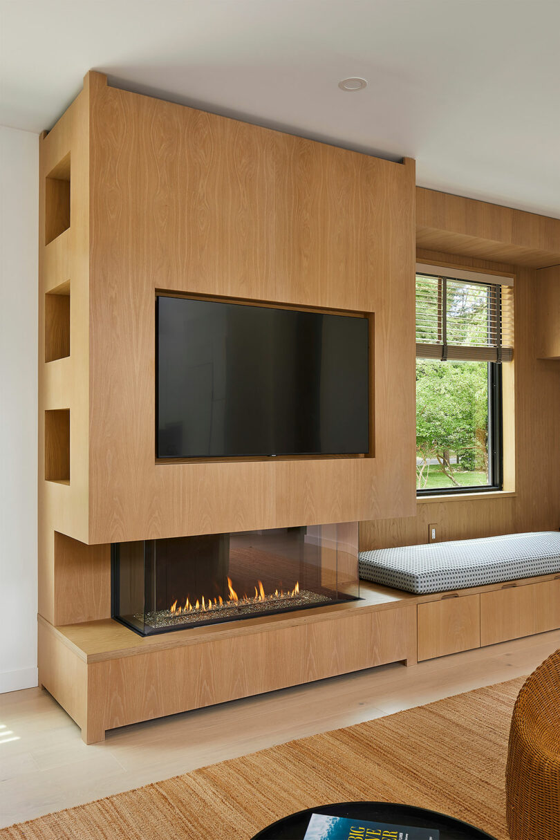 The Shift House modern living room boasts light wood paneling, a built-in fireplace, and a flat-screen TV. A window with a bench seat delicately bathes the space in natural light.
