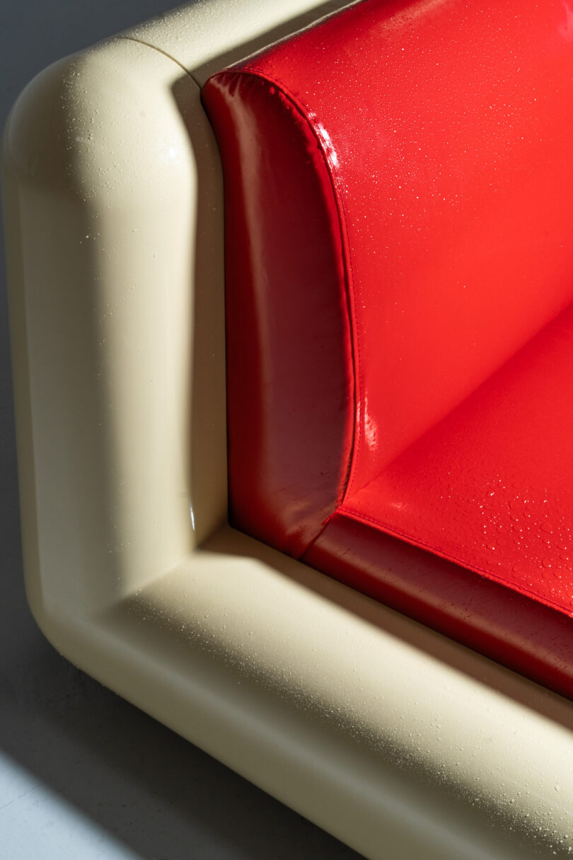 Close-up of a corner of a sofa with glossy red cushions and a cream-colored frame. The surface appears to have moisture on it, possibly indicating wetness