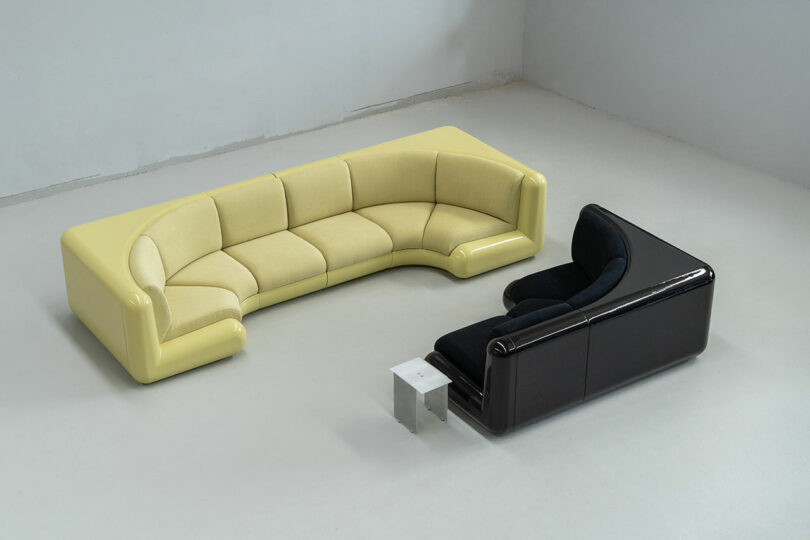 Curved yellow sofa and smaller curved black sofa arranged around a small square white table in a minimalist, light-gray room.
