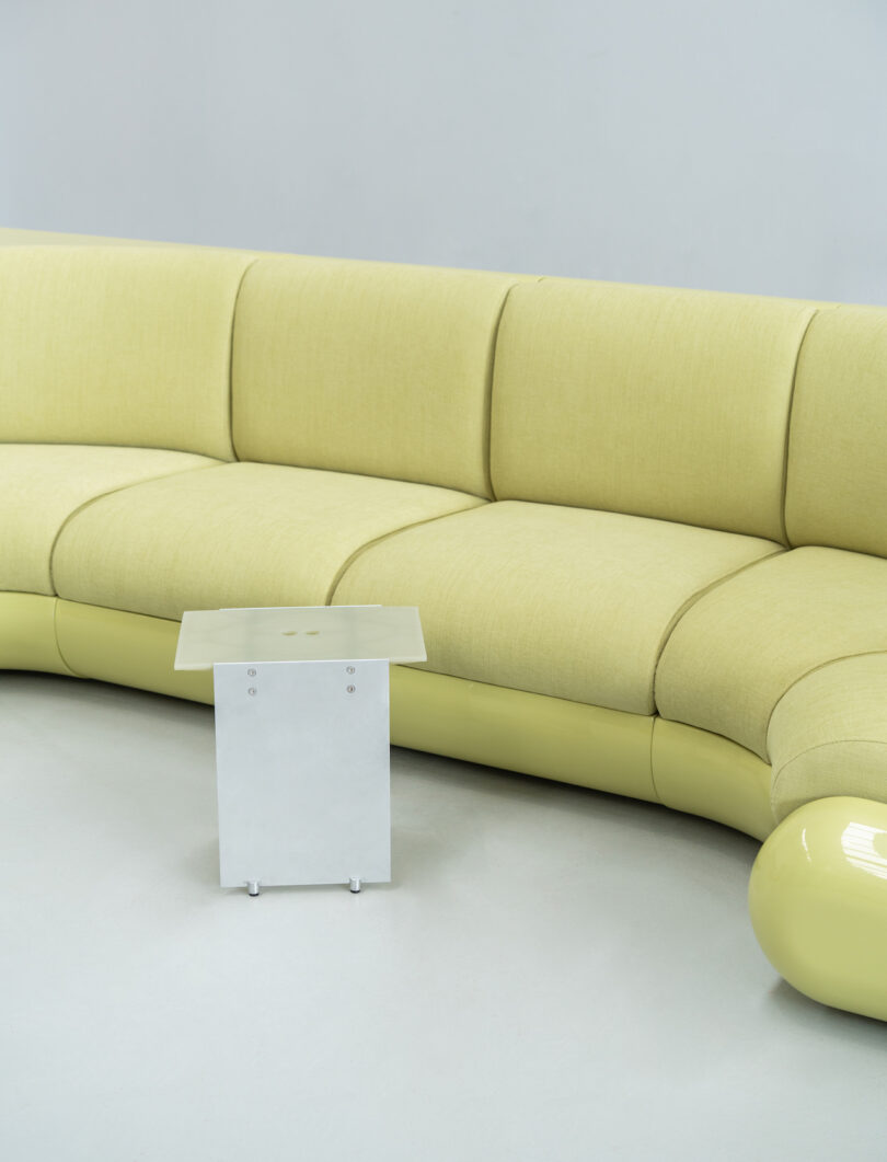 A pale yellow, curved sofa is positioned against a light gray wall. In front of it, a small, white, rectangular side table is placed on the floor