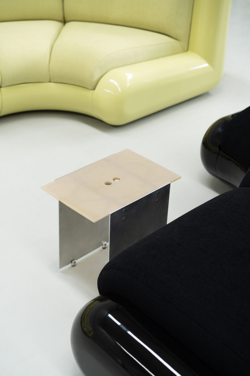 A small rectangular table with a light surface and dark legs is placed between a light yellow curved sofa and a black cushioned seat