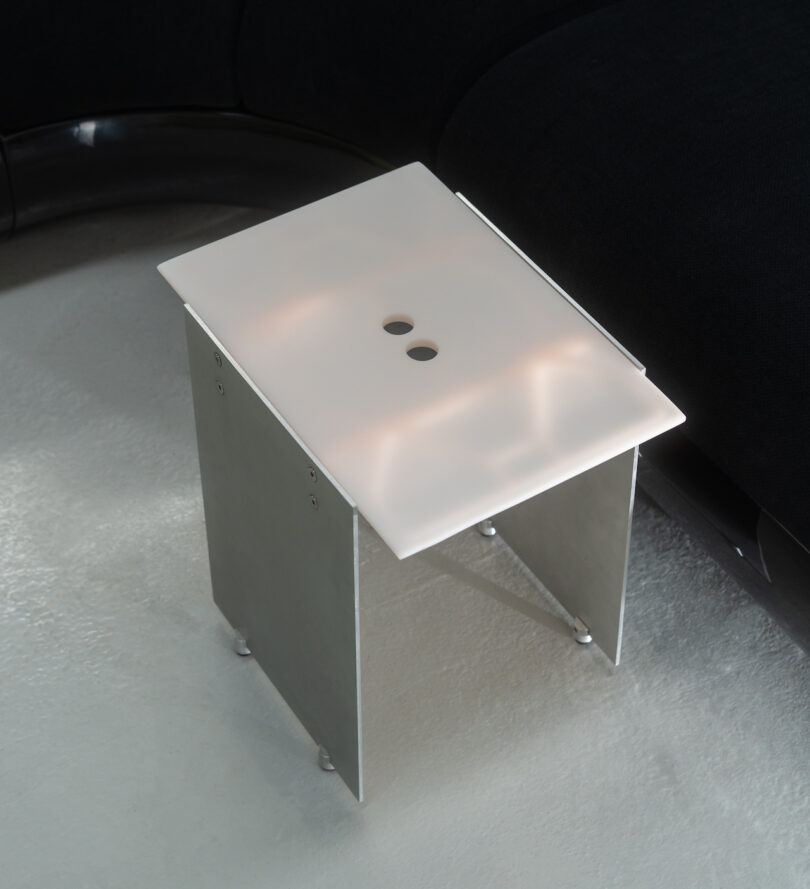 A modern, minimalist side table with a frosted glass top featuring two circular cutouts, positioned on a light-colored floor next to a dark sectional sofa