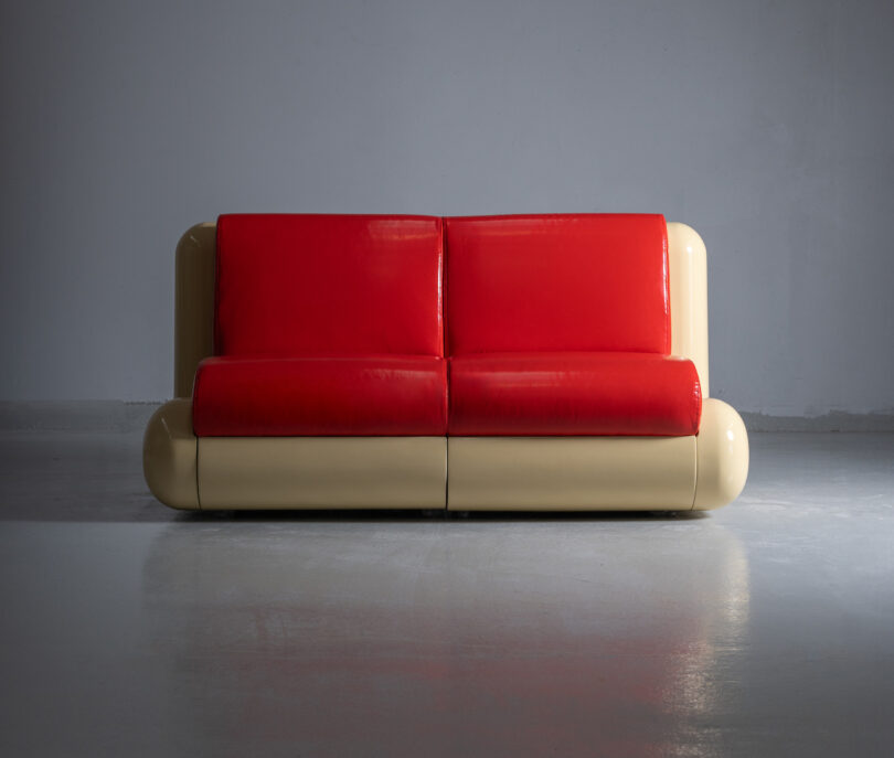 A modern sofa with two red cushions and a beige frame set against a plain gray background