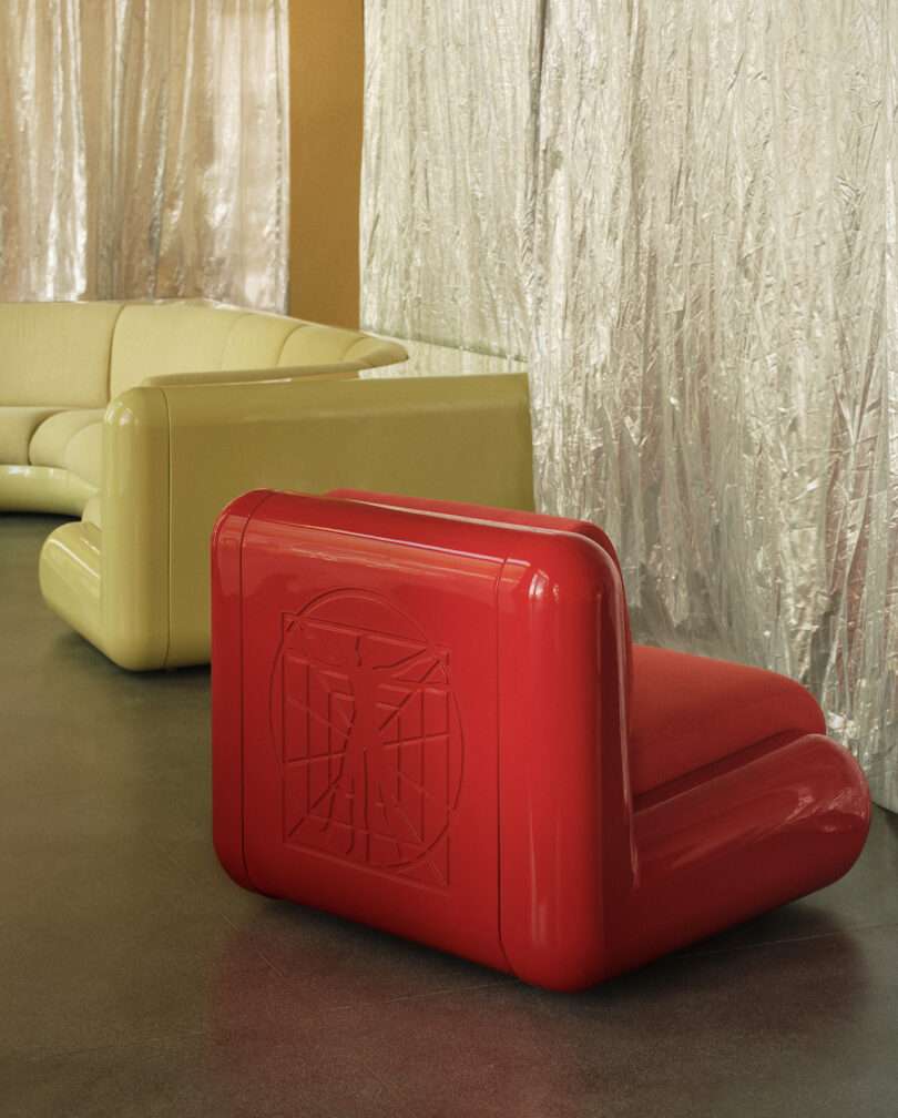 Red chair with geometric design next to a cream-colored sectional sofa on a smooth floor, set against metallic curtains
