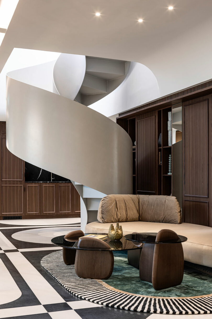 A modern living area with a tan sofa, round glass coffee table, dark wood accents, and a large spiral staircase in the background.