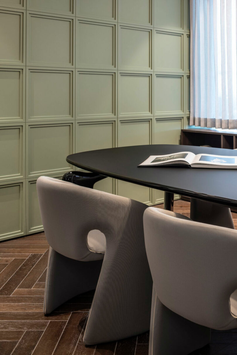 A modern conference room with a black oval table, two gray chairs, an open book, wood herringbone flooring, and a green paneled wall.