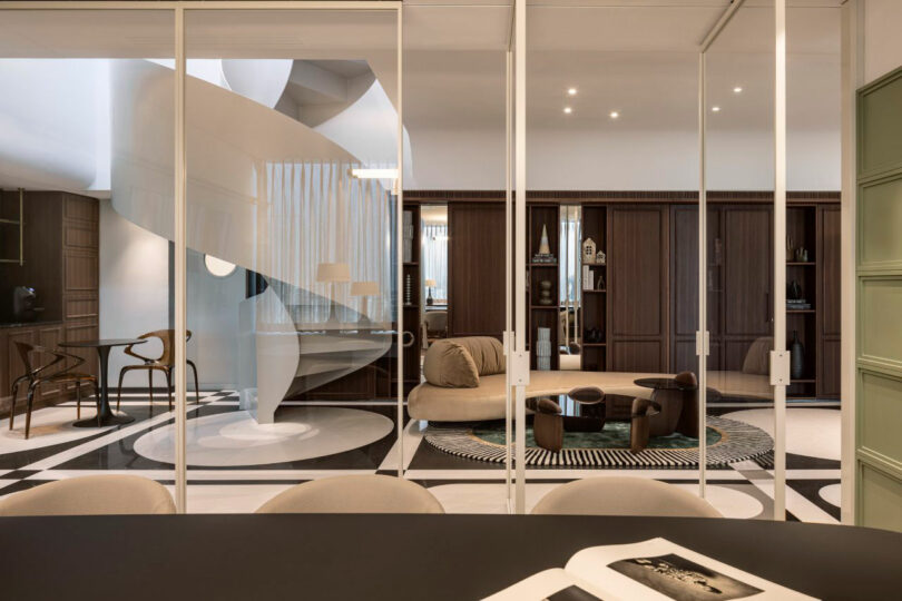 Modern living room viewed through glass walls, featuring a spiral staircase, round sofa, built-in wooden shelves, and a circular rug on a patterned floor.