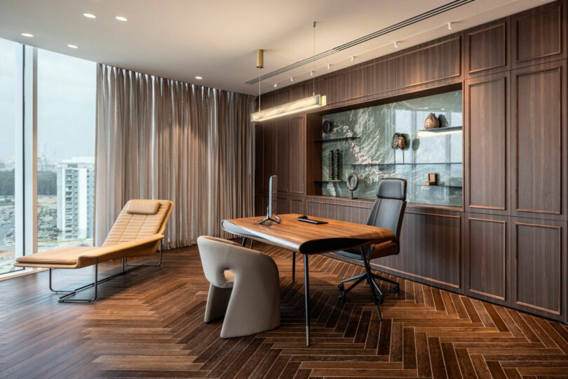 Modern office with a wooden desk, two chairs, a lounge chair, built-in shelves, wood herringbone floor, large window, and neutral-toned decor.