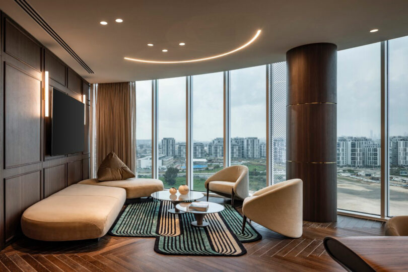 Modern living room with beige furniture, round chairs, a large TV, and floor-to-ceiling windows showing a cityscape with apartment buildings.