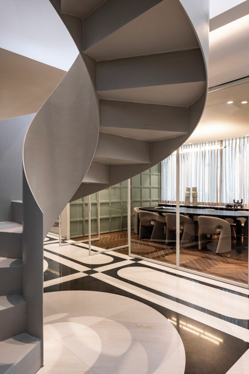 A modern interior featuring a gray spiral staircase, geometric black-and-white flooring, and a dining area with a long table and chairs near large windows with sheer curtains.