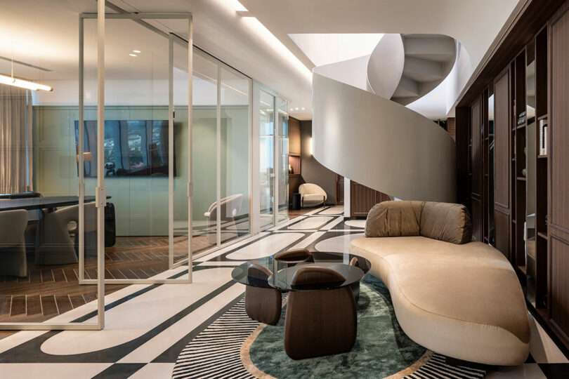 Modern living room with a curved beige sofa, glass coffee table, spiral staircase, patterned floor, built-in shelves, and glass wall separating an office area.