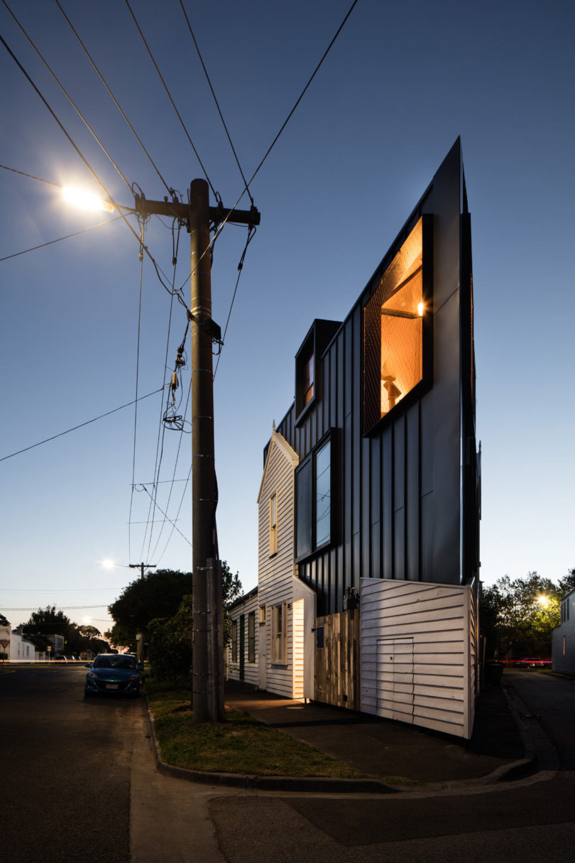 Modern triangular-shaped house with black and white exterior panels, large windows, and sharp roofline at dusk on a quiet residential street with a streetlight and parked car.