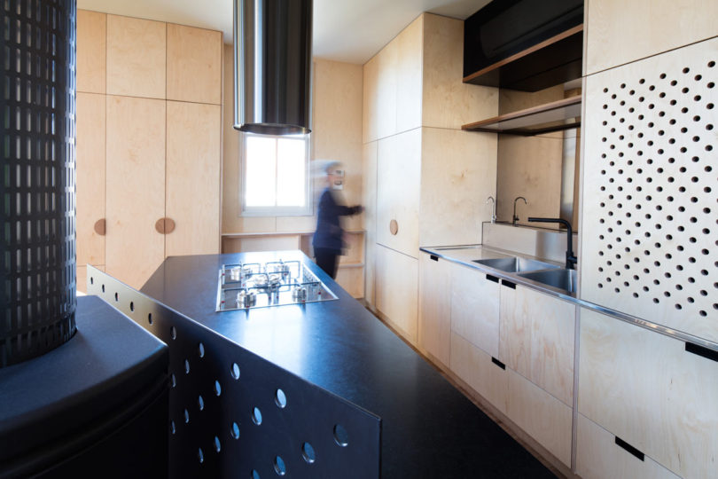 Modern kitchen inside a triangular-shaped house with light wood cabinetry and black countertops, featuring perforated cabinet fronts and an island with a built-in gas stove; a blurred person stands near the window.