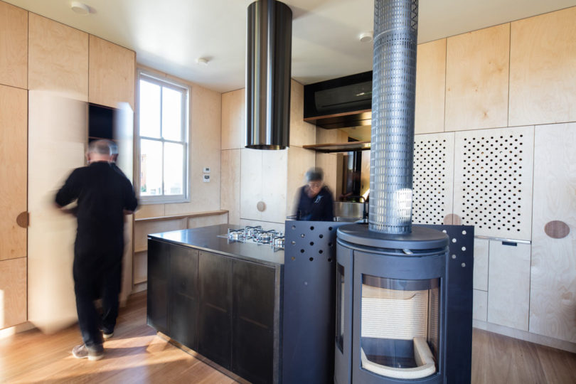 Modern kitchen with light wood cabinets in a triangular-shaped house, featuring a black island with a built-in gas cooktop, a cylindrical stove, and two blurred people in motion.