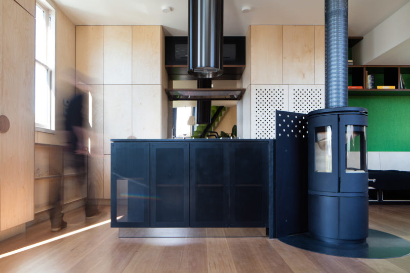 Modern kitchen in a triangular-shaped house with light wood cabinets, a black island, a cylindrical wood stove, and a blurred person walking on the left.