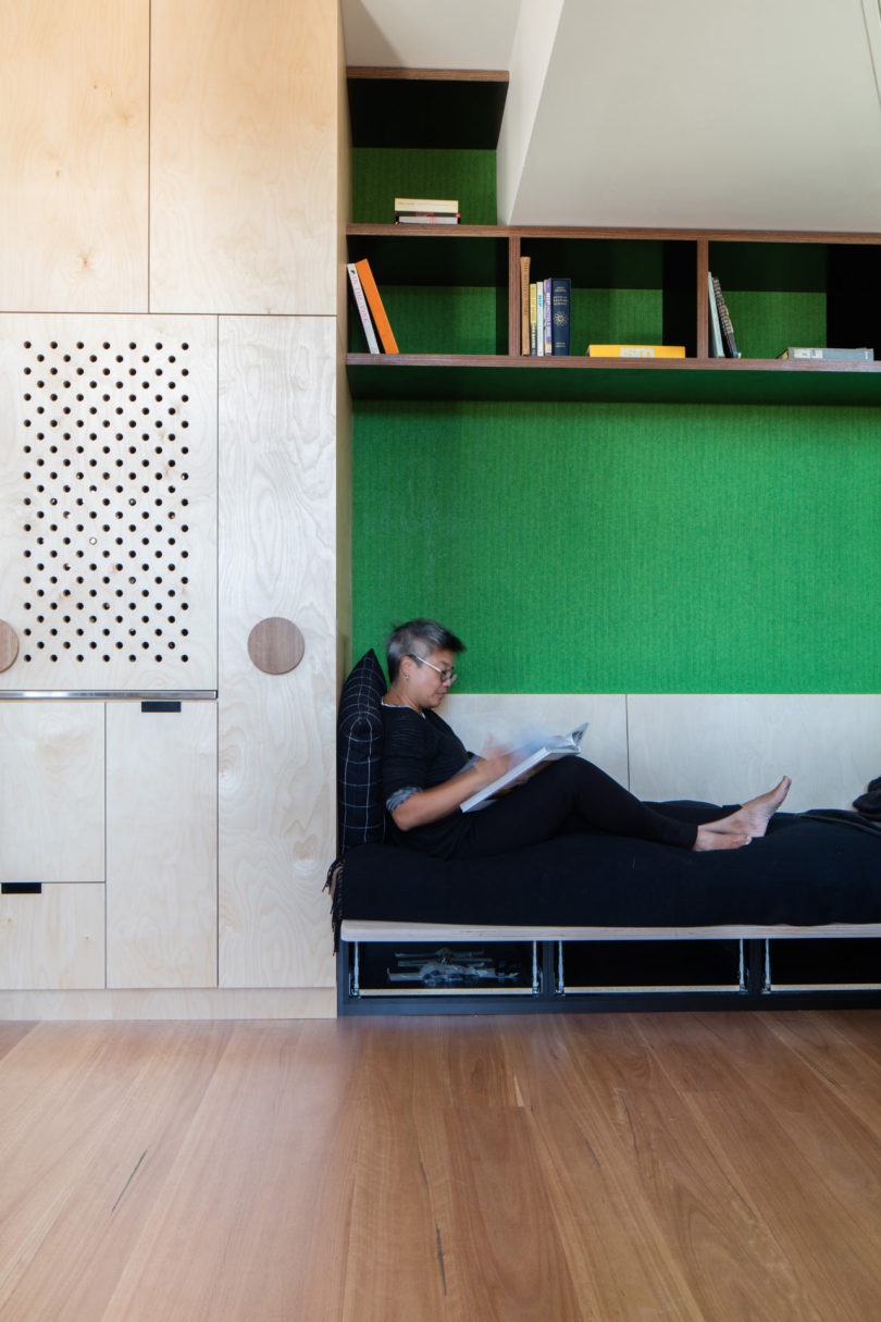 A person sits cross-legged on a built-in daybed, reading a magazine in a modern, triangular-shaped house with green wall panels and wooden shelves.