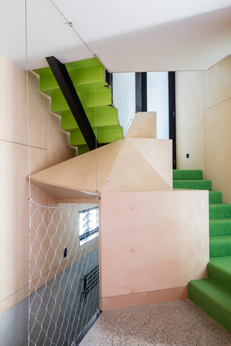 A modern staircase in a triangular-shaped house features bright green steps, light wood paneling, a metal safety net, and terrazzo flooring within a minimalist interior.