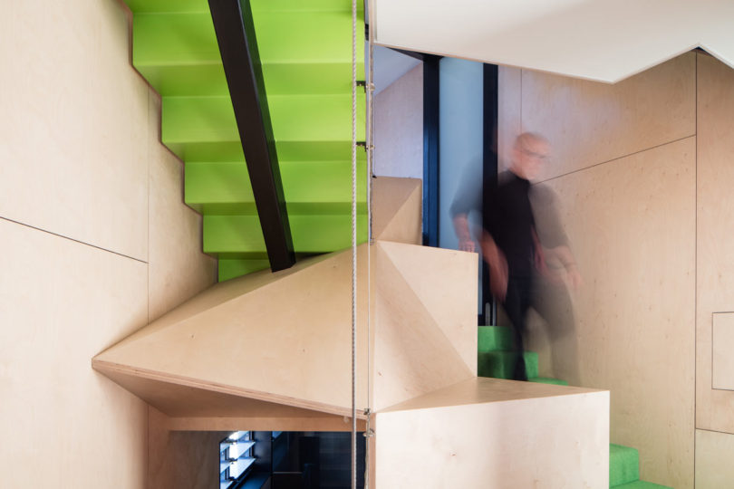 A person descends a modern staircase with bright green steps and light wood paneling in a triangular-shaped house, featuring geometric design elements and a blurred motion effect.
