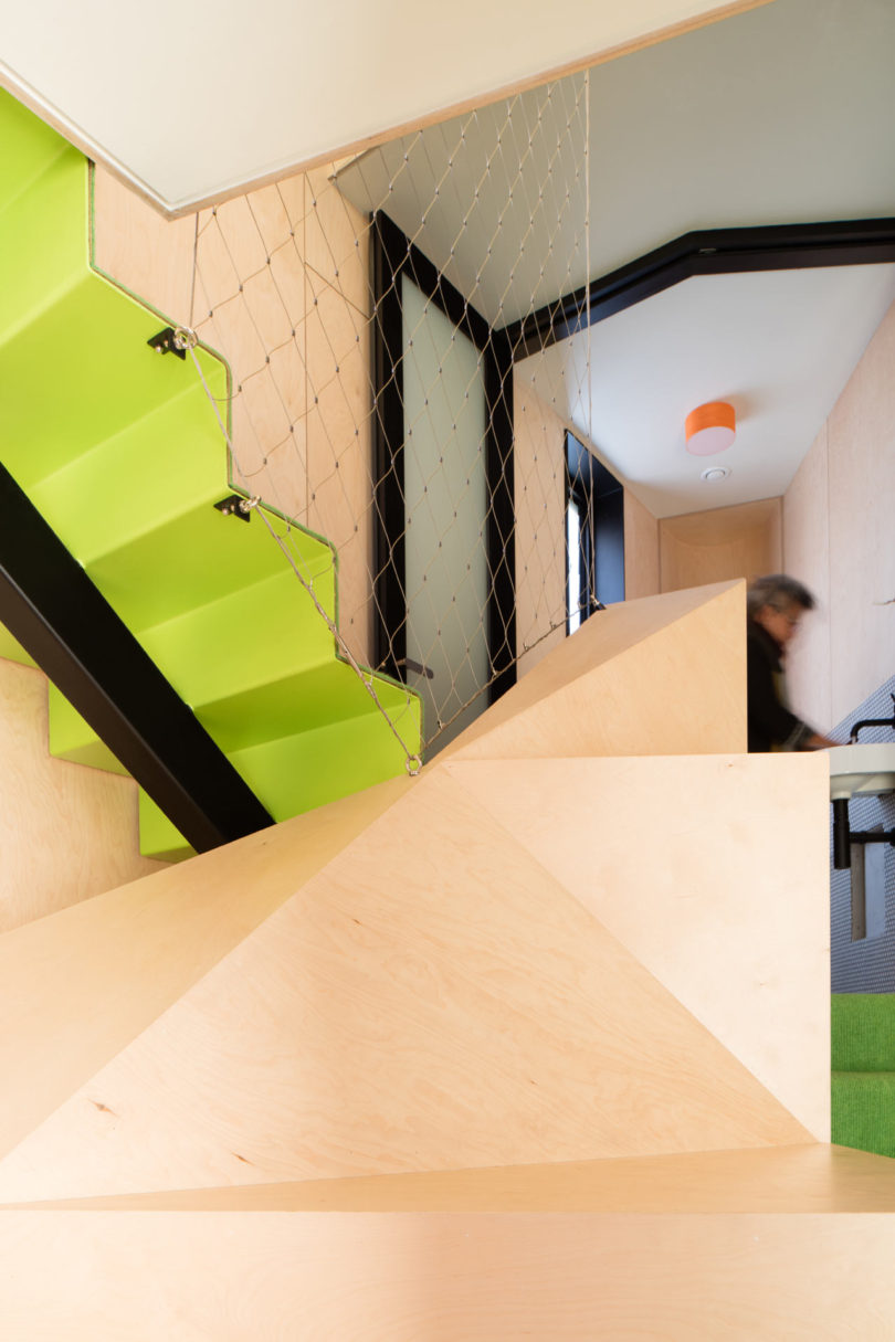 Modern interior of a triangular-shaped house with light wood geometric walls, a bright green staircase featuring a metal net railing, and a blurred person walking in the background.