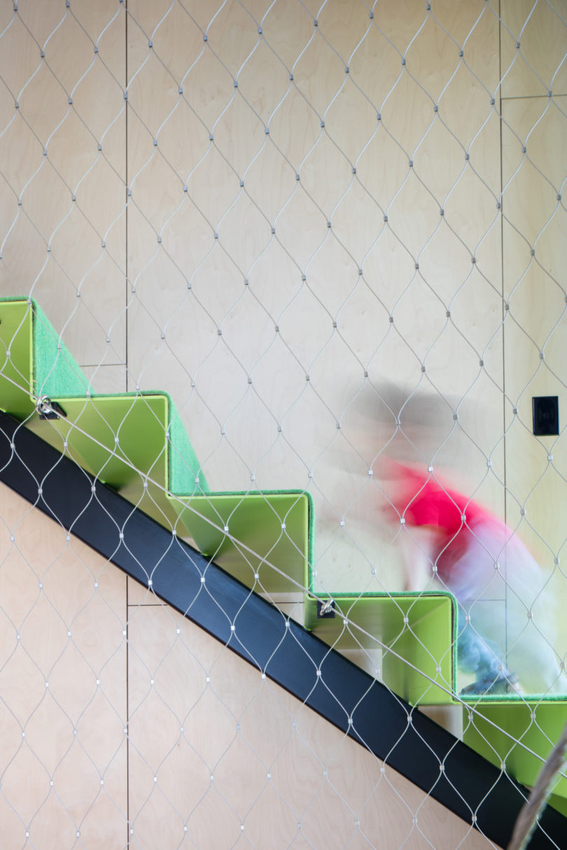 A blurred figure ascends a zigzag-shaped green staircase behind a wire mesh, framed by light wood panel walls in the background of a triangular-shaped house.