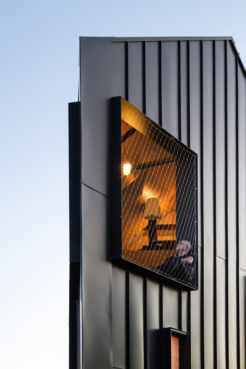 A modern, triangular-shaped house with dark metal siding features a protruding window box with mesh and a person inside, illuminated by a hanging light bulb.