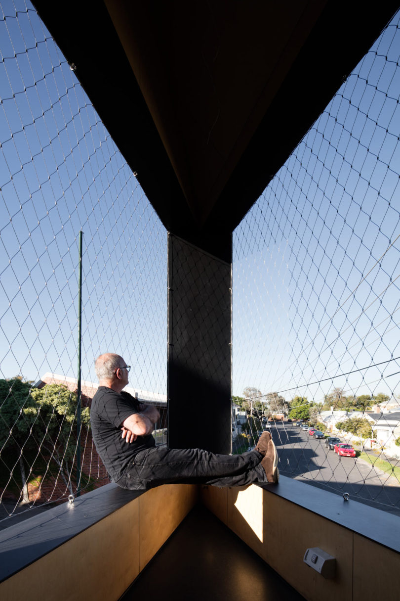 A person sits on a window ledge in a modern building, looking outside through mesh-protected glass at a sunny street and neighborhood featuring a distinctive triangular-shaped house.