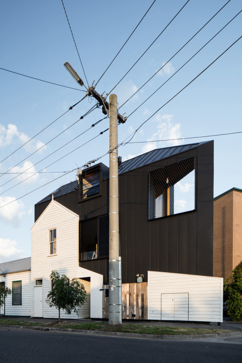 A modern black building with large windows rises behind a smaller, triangular-shaped white house, with a utility pole and power lines in the foreground.