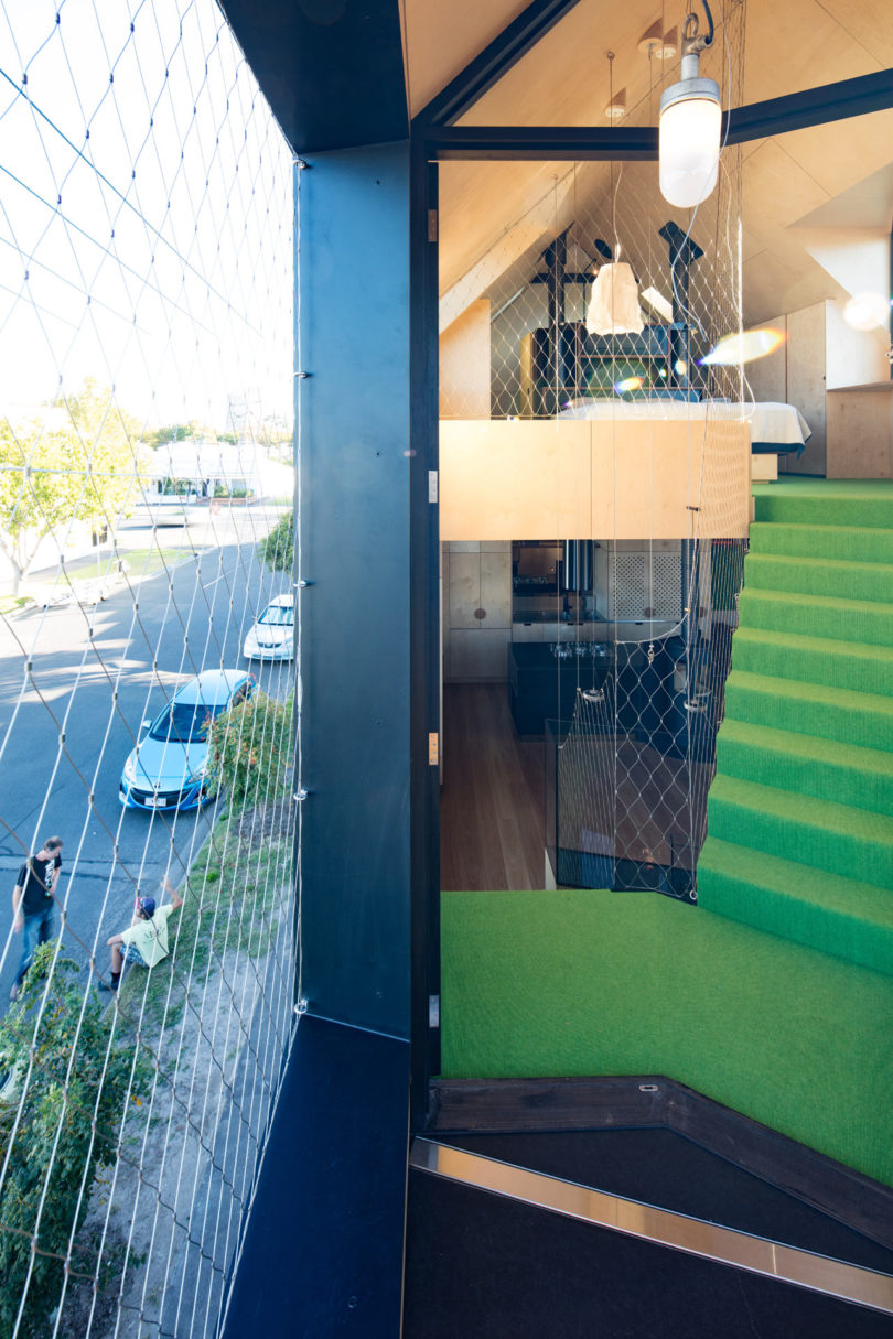 A modern triangular-shaped house interior with green carpeted stairs, wood-paneled walls, and large glass windows overlooking a street with parked cars and pedestrians.