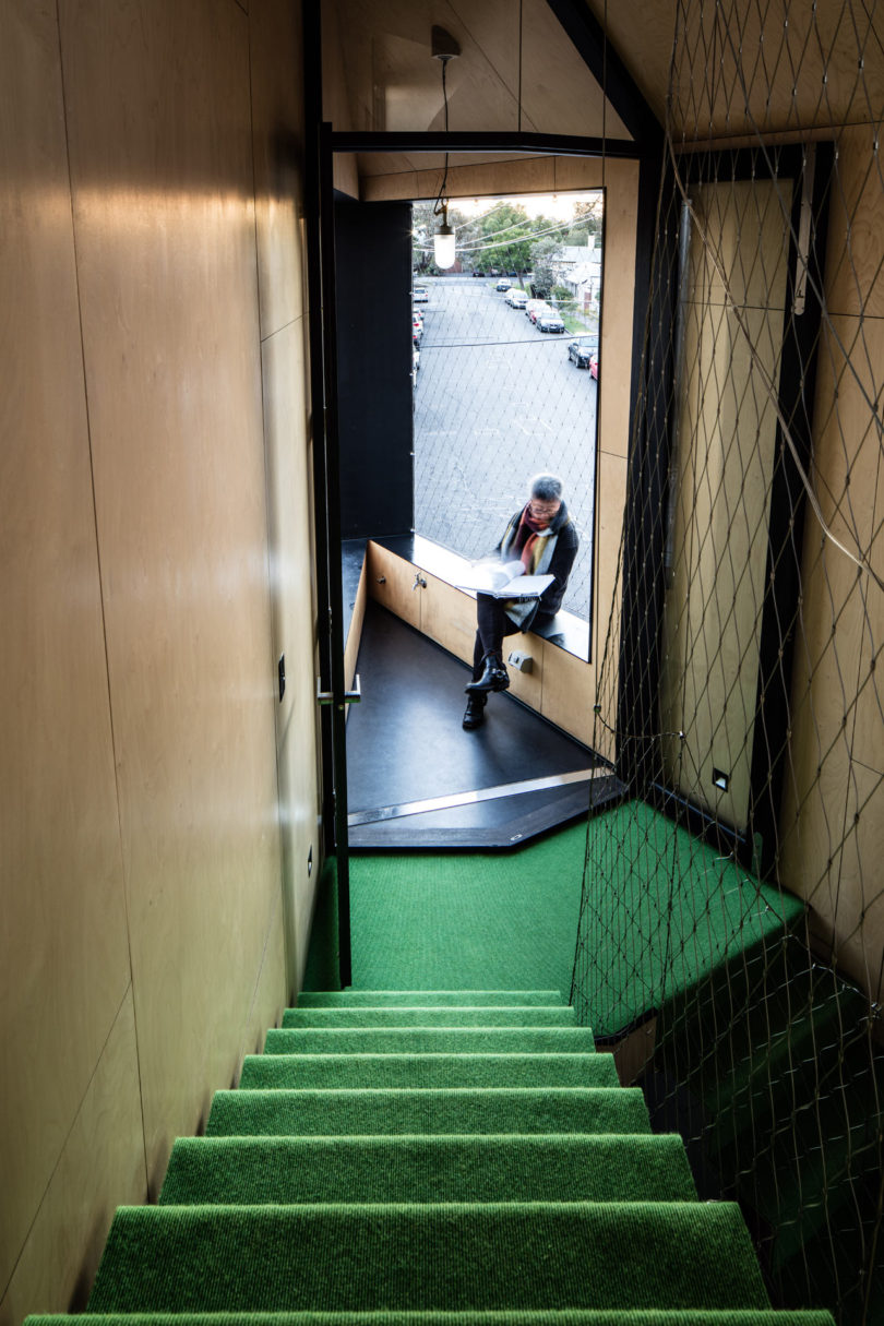 A person sits reading by a window at the end of a hallway in a triangular-shaped house, with green carpeted stairs and wood-paneled walls, overlooking a street with parked cars outside.