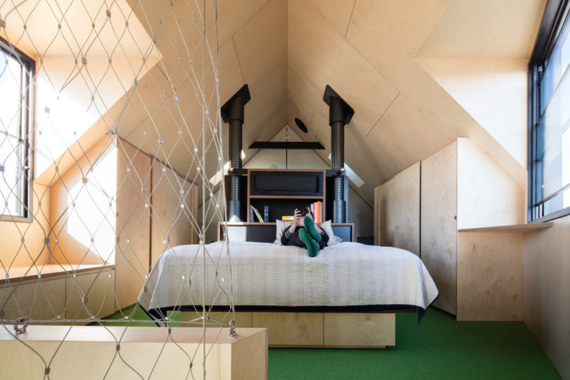 Person lying on a bed in a modern attic bedroom inside a triangular-shaped house, featuring angled wooden walls, green carpet, large windows, built-in storage, and a safety net along one side.