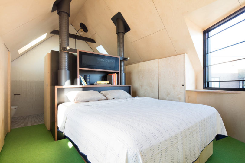 A modern attic bedroom in a triangular-shaped house, featuring sloped ceilings, a double bed, built-in shelves, two vertical vents, and a large window that fills the space with natural light.