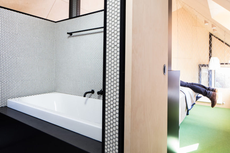 A modern bathroom with a white bathtub and black fixtures is separated by a partial wall from a bedroom, all nestled within a striking triangular-shaped house, where a person is lying on the bed with their legs visible.