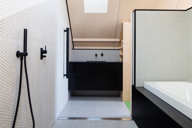 Modern bathroom inside a triangular-shaped house, featuring white hexagonal tile walls and floor, black fixtures, black vanity, walk-in shower, and bathtub. Light wood accents on the ceiling and wall add warmth to the unique space.
