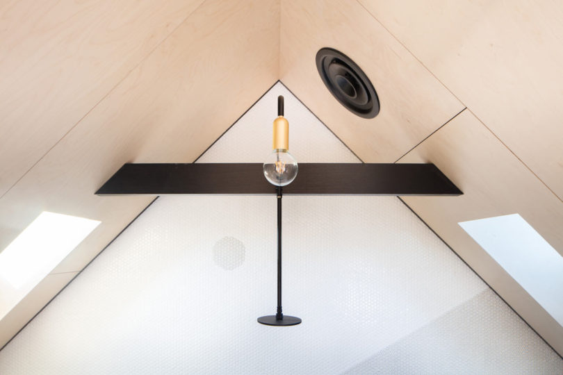 Modern light fixture with exposed bulb and black horizontal bar mounted on a sloped ceiling with light wood panels and skylights, ideal for a triangular-shaped house.