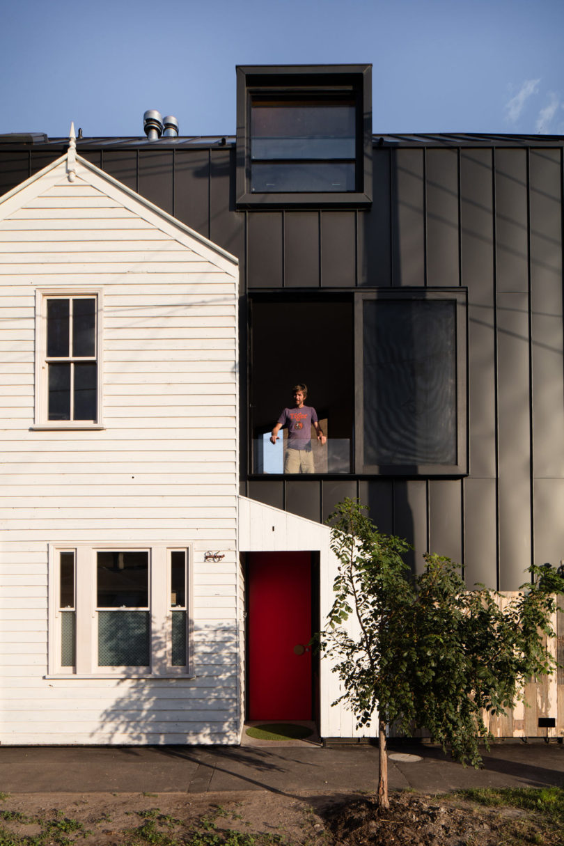 A person stands on a recessed balcony of a modern black, triangular-shaped house attached to an older white house, with a red front door and a small tree in front.