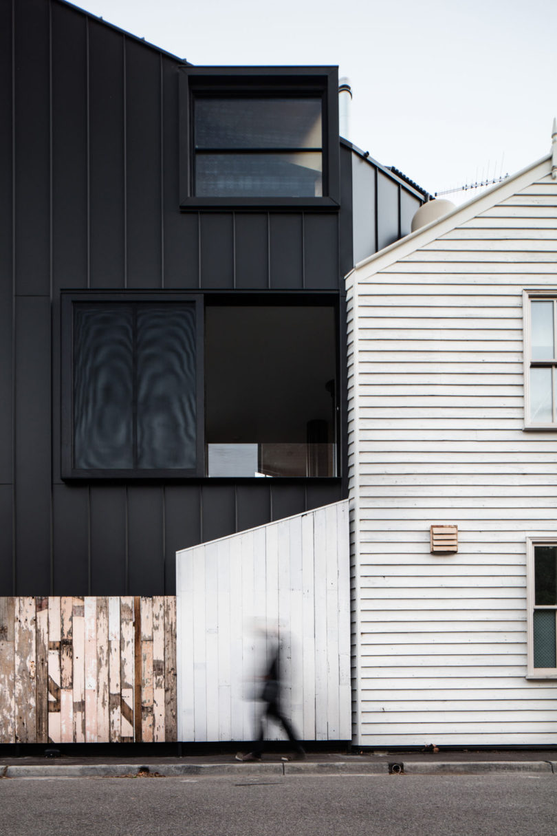 A person walks past two modern houses—one a striking triangular-shaped house with black cladding and large windows, the other featuring white siding—separated by a slanted fence.