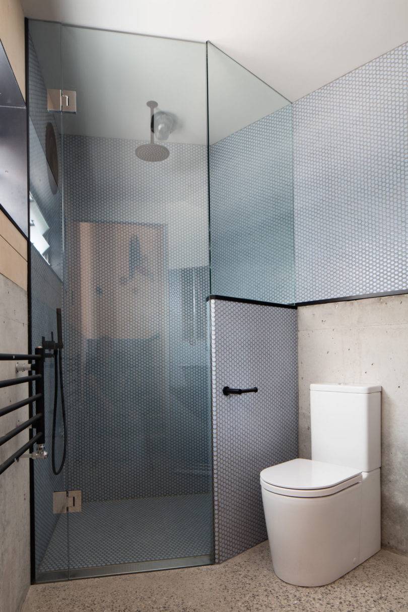 Modern bathroom in a triangular-shaped house, featuring a glass-enclosed shower, wall-mounted showerhead, heated towel rack, and a white toilet on a terrazzo floor beside concrete and tiled walls.