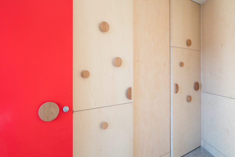 A red door with a round wood handle sits beside light wood cabinets featuring large circular wooden knobs in a minimalist, triangular-shaped house interior.