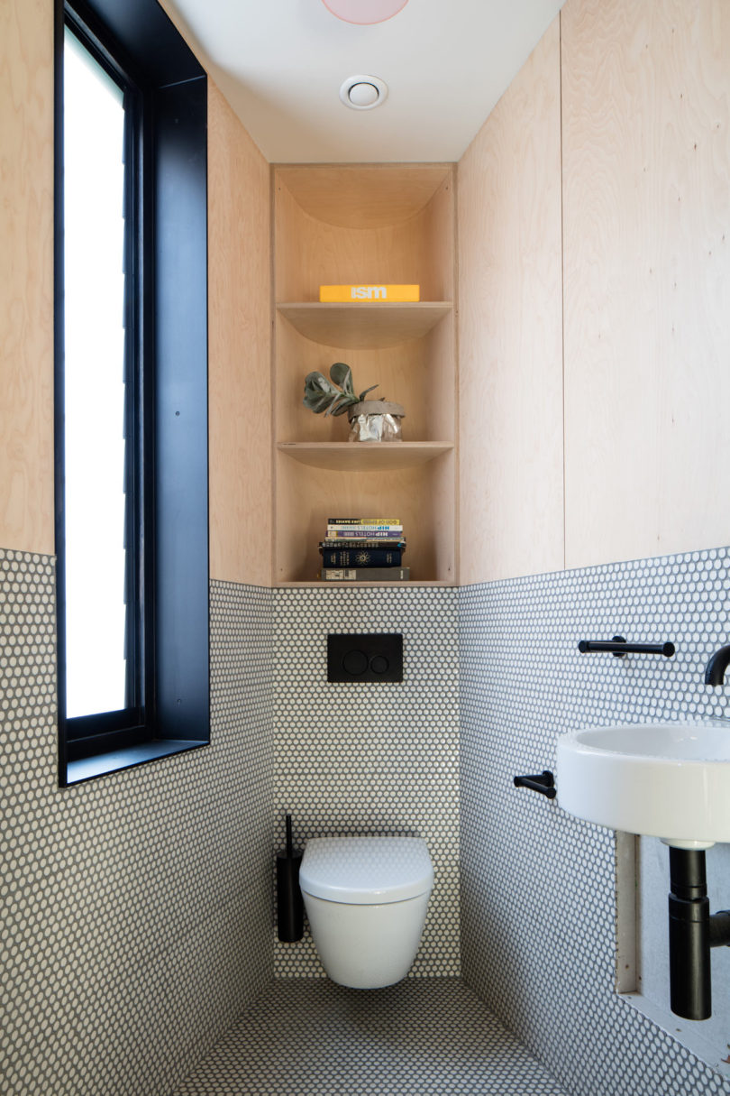 A modern bathroom in a triangular-shaped house with a wall-mounted toilet, small white sink, built-in shelving above the toilet, and round tile walls and floor.
