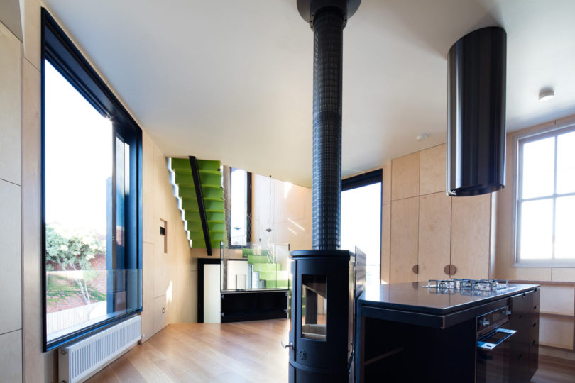 Modern kitchen inside a triangular-shaped house, featuring light wood paneling, a black stove and island, large windows, and a green staircase in the background.