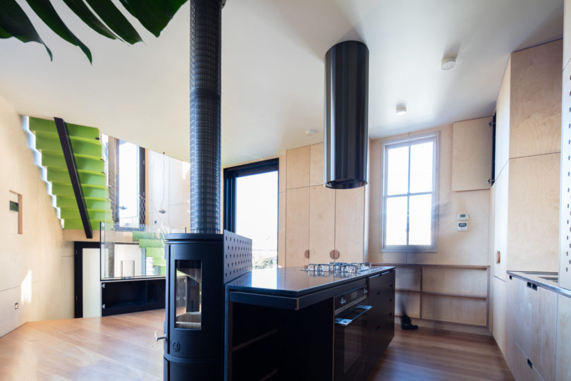 Modern kitchen with black island, central stove, light wood cabinetry, and a large window, all set within a triangular-shaped house featuring a striking green staircase to the upper floor.