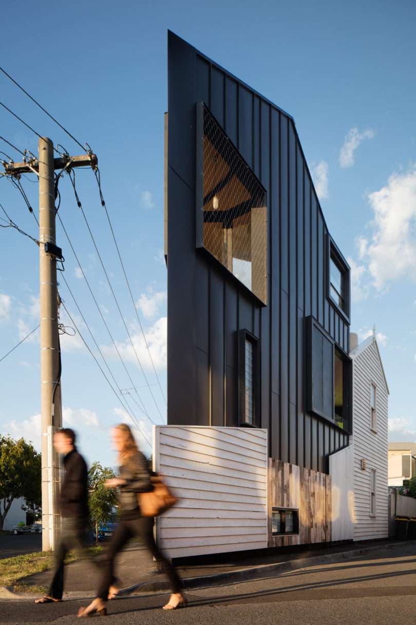 Two people walk past a modern, triangular-shaped house with angular black exterior, large windows, and a white fence under a clear sky.